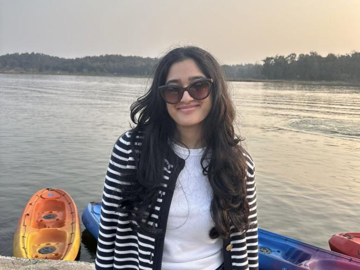 Urja Mehta standing in front of a body of water with kayaks behind her.